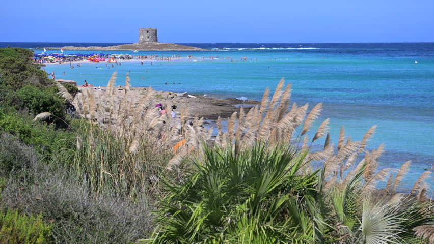People enjoy the beach while others relax on the sand near a historic tower by the sea. Sardinia, Italy