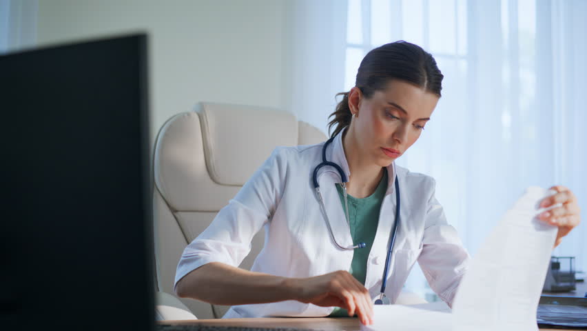 Serious therapist examining diagnosis papers at light office closeup. Focused doctor check radiograph scan successful treatment working computer. Healthcare professional analyzing in medical clinic