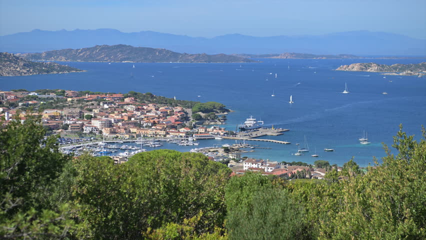 A view shows Porto Rotondo in Italy with boats in the water and hills in the background on a clear day.