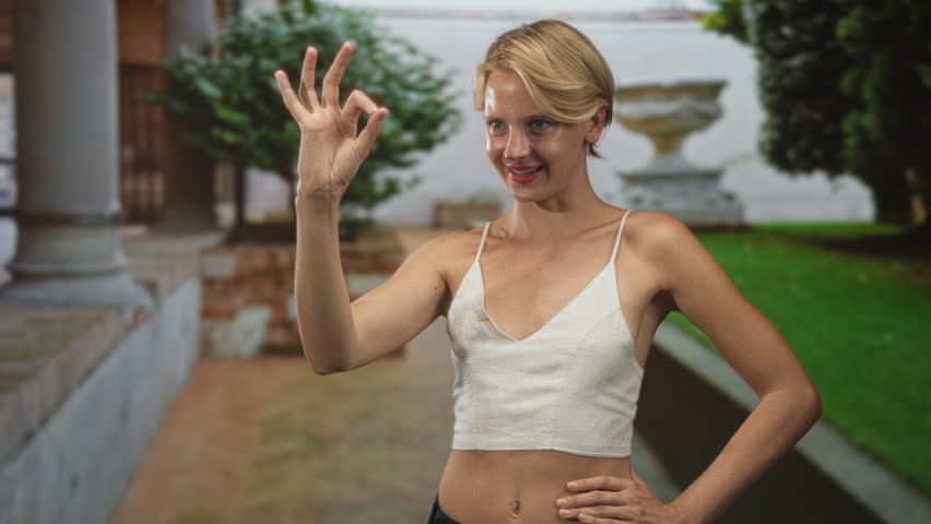 Woman making ok sign with right hand, white cami and visible midriff, smiling in building courtyard with classical urn and columns; confidence ease.