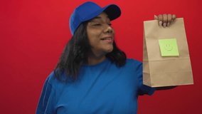 African american courier woman smiling holds paper bag in red studio with blue cap and uniform; friendly service. - Powered by Shutterstock - Get 15% off with code: PIKWIZARD15