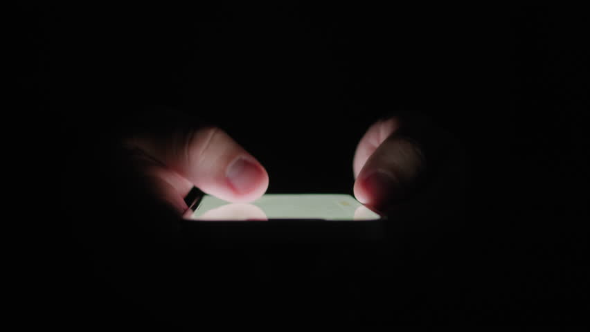 Phone at night, Close-up macro hands of a man scroll news feed on mobile phone app application. Screen of the mobile phone illuminates male hand. 