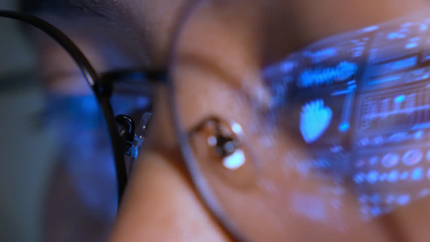 A person wearing glasses uses a laptop, with a real-time digital medical interface reflected in the lenses, representing healthcare analytics, clinical monitoring, and advanced medical technology.
