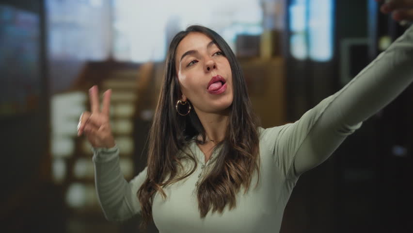 Young hispanic woman flashing peace sign and sticking out tongue with smartphone in hotel building; youthful joy.