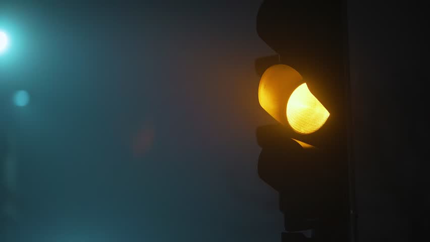 Close-up shot of a blinking yellow traffic light glowing through dense fog at night. The warm light cuts through the misty atmosphere