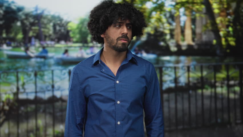 Man with curly hair in blue shirt looking to the side in park by an ornamental iron fence and boating lake, surrounded by green trees and a walking path; quiet concern.