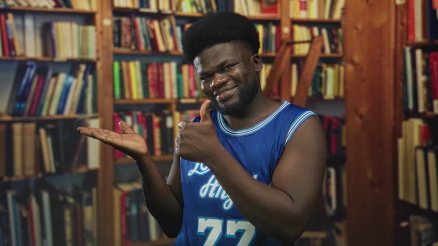 Man pointing finger to palm in building library aisle, open hand visible and smiling while wearing blue basketball jersey; playful confidence.