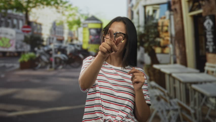 Woman points finger at camera showing fingers and smiling on street cafe terrace with glasses and striped shirt; playful confidence.