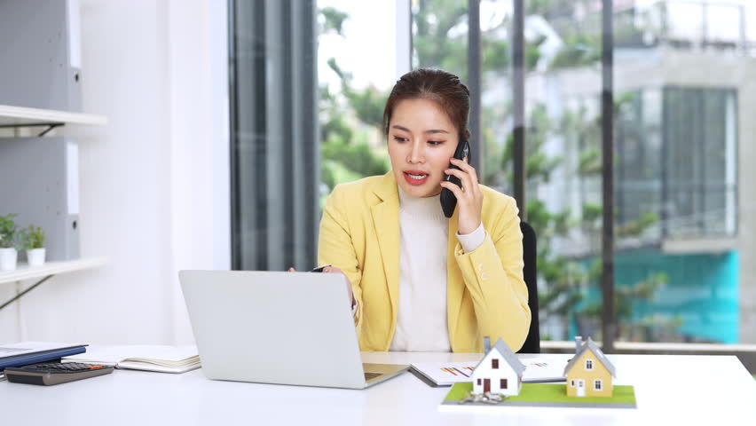 Confident asian business lady, broker real estate agent or manager, sitting at table in a modern office, working with laptop, talking to client or employees on smartphone, smiling.