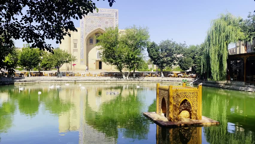 Lyabi-Hauz ensemble in the heart of Bukhara, Uzbekistan. The shot features the tranquil central pond reflecting the ancient mulberry trees and the ornate facade of the Nadir Divan-Begi Madrasah