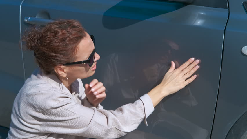 Woman examining scratches on car door. Woman wearing dark sunglasses carefully inspecting gray car exterior, scanning surface for potential scratches or dents under bright sunlight