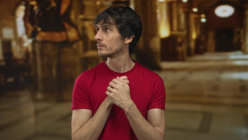 Man in red shirt holds clasped hands beneath vaulted arches in stone church interior with golden light from stained glass windows; devotion.