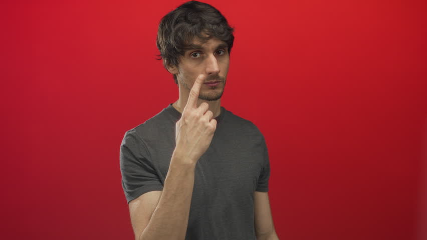Young man in grey tshirt showing thoughtful gaze while pointing finger to cheek in a red studio; insight contemplation.