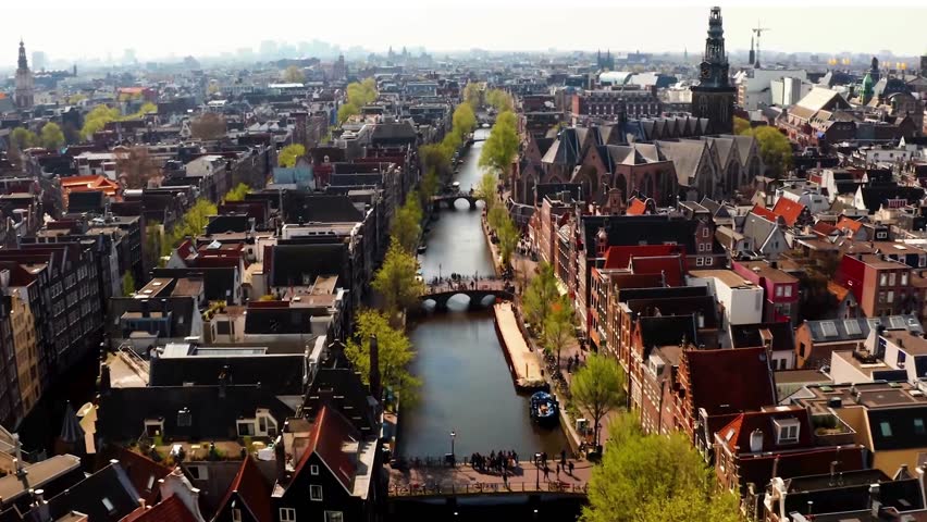 Aerial drone view of Amsterdam, Netherlands, showing the cityscape with historic canals, urban architecture, and bustling streets. Scenic overview of the Dutch capital, highlighting iconic buildings, waterways, and vibrant city life from above. Perfect for travel, tourism, and urban landscape visuals