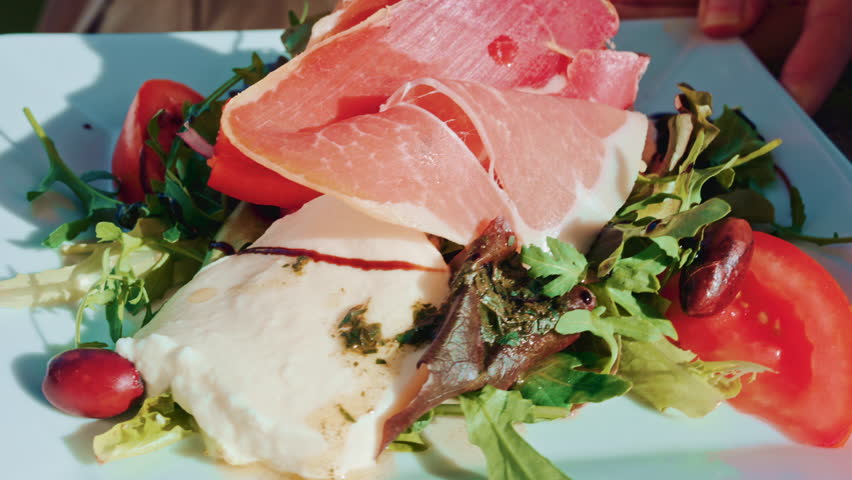 Close up of a gourmet salad with fresh mozzarella, cured meat slices, tomatoes, olives and mixed greens on a white plate in bright daylight