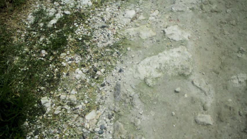 This is a close-up view of a dusty, gray dirt path showing its rough texture with scattered dry grass and numerous loose stones on the edges, suggesting a rural or unpaved road