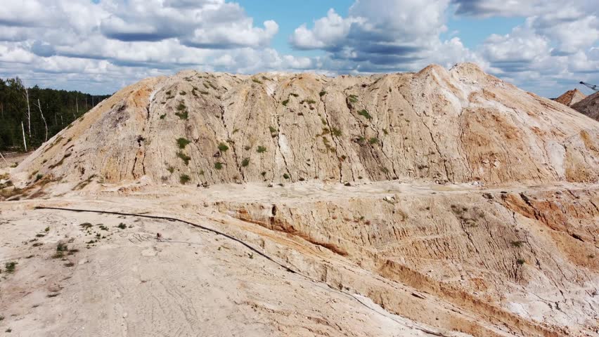 Waste clay and rock dumps on the ilmenite quarry edge against the cloudy sky, aerial view while moving forward in summer sunny day

