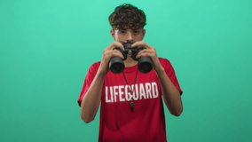 Teenage lifeguard in red shirt holds binoculars to eyes with mouth slightly open and wears metal whistle necklace in studio; vigilance duty. - Powered by Shutterstock - Get 15% off with code: PIKWIZARD15