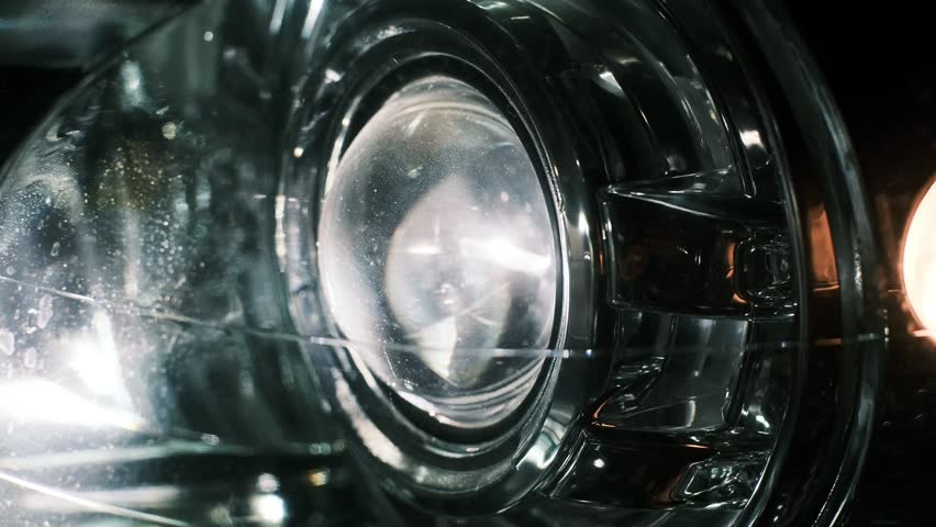 Close-up of a car headlight shining brightly at night, showcasing reflective glass, LED beams, and turn signal illumination. Dramatic automotive lighting detail in a dark environment.