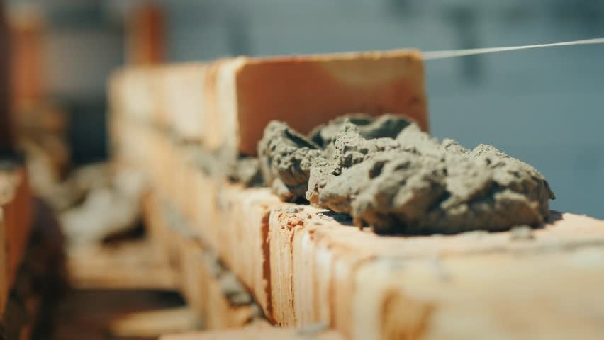 This high-quality 4K video provides a detailed look at the fundamental craft of bricklaying, a cornerstone of global construction and architecture. The footage captures a skilled mason precisely placing individual bricks onto a prepared bed of mortar, showcasing the rhythmic and technical nature of the trade. The bricks, likely made from kiln-fired clay, are aligned with careful attention to level and plumb, ensuring the structural integrity of the rising wall. The mortar—a vital mixture of cement, sand, and lime—is seen being spread and trimmed, creating the necessary bond between the masonry units.

The video highlights the material science behind modern building, where the strength of the cementitious bond meets the durability of the clay brick. This process is essential for creating everything from residential homes to massive industrial structures. The close-up shots allow viewers to see the texture of the wet mortar and the porous surface of the bricks, emphasizing the tactile elements of the building trade. Whether for educational purposes or industrial documentation, this sequence serves as a perfect visual representation of manual labor, craftsmanship, and civil engineering in action, illustrating how individual components are transformed into a solid, lasting structure through expert skill.