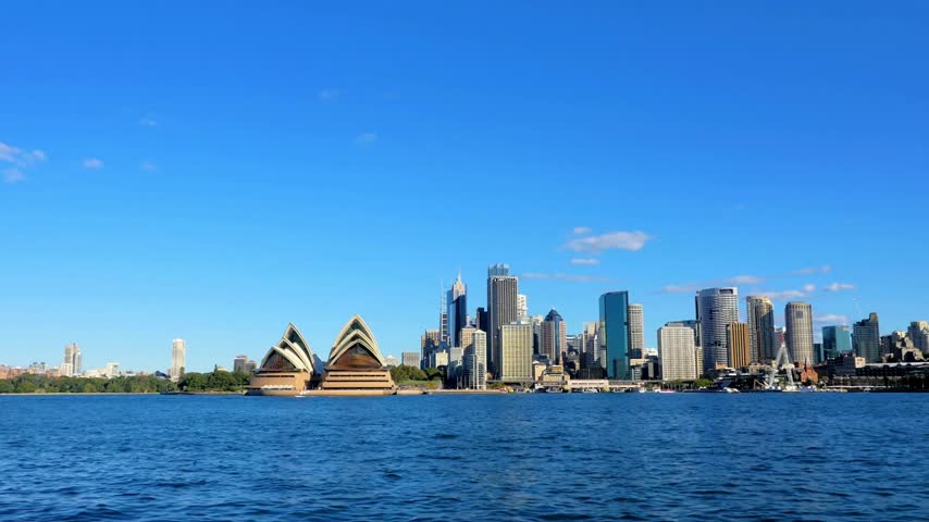This sweeping 4K view captures the iconic skyline of Sydney, Australia, showcasing the city’s unique blend of modern high-rises and historic landmarks. Dominating the foreground is the Sydney Opera House, a UNESCO World Heritage site world-renowned for its distinctive white "shell" or "sail" design. As a global symbol of Australian culture and architectural innovation, the Opera House sits majestically on the edge of the blue harbor waters. The footage highlights the vibrant Central Business District (CBD), with its towering skyscrapers reaching toward the horizon. This panoramic perspective emphasizes Sydney