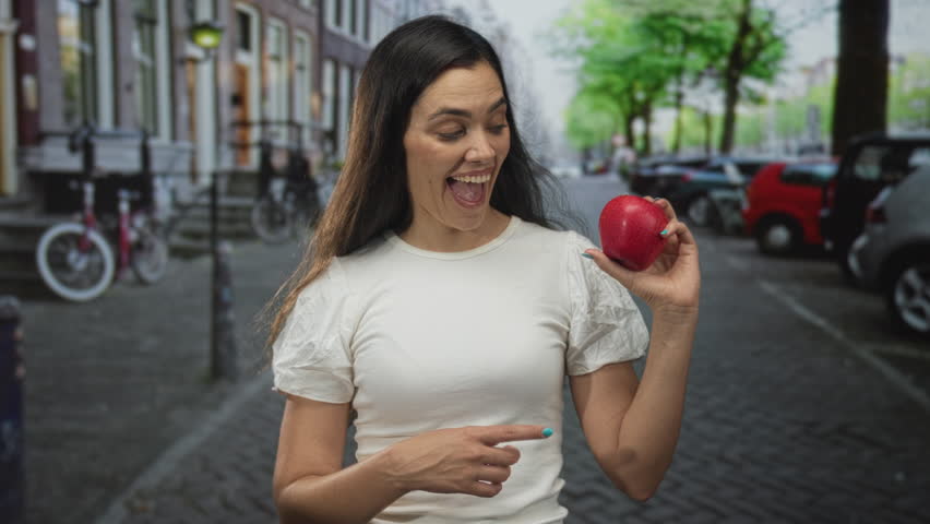 Young hispanic woman holds red apple in one hand while gazing at it on a cobblestone street lined with parked cars and bicycles; doubt.