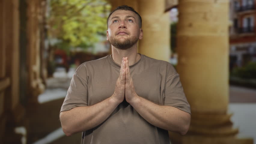 Man with bare hands pressed together in prayer under stone columns on an old town street looking upward; quiet faith.