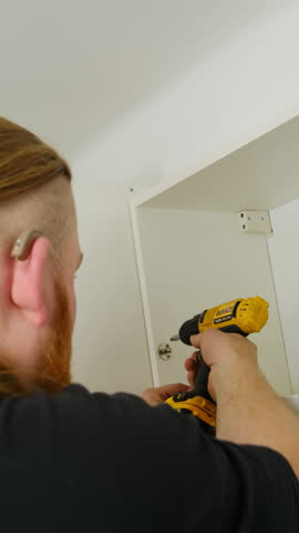 Handyman with electric screwdriver takes small screws from shelf of cabinet closeup. Bearded man assembles domestic furniture manually with tool at home vertical shot