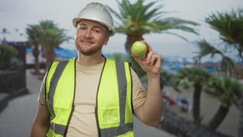 Man engineer in hardhat and hi vis vest finger pointing while holding apple on street by palm trees near seaside; safety confidence.