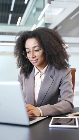 Portrait of smiling African American businesswoman sitting at desk at workplace in business office. Happy female manager in glasses and jacket works on computer and looking at camera. Vertical video