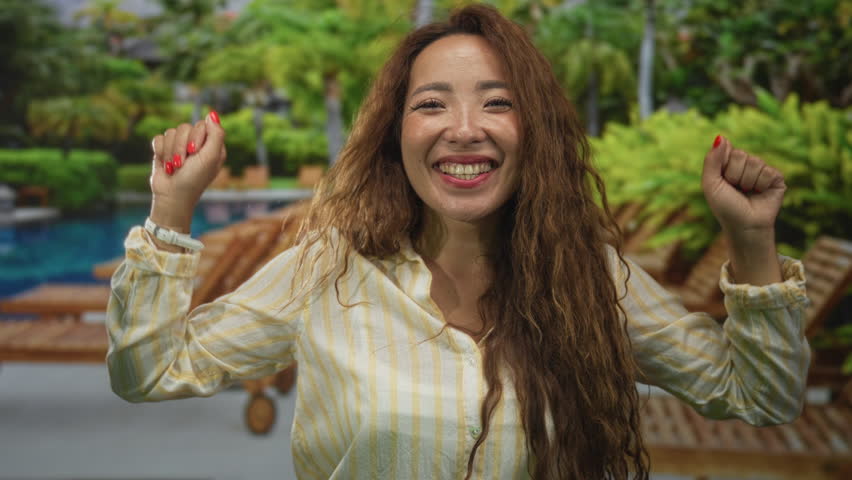 Woman smiling raises arms at hotel pool area wearing striped shirt showing face and hands in outdoor resort with sunbeds and palms; joy vacation celebration.