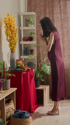 Vertical shot of young Asian woman having prayer by home altar with vase, fruits, and burning incense stick during Tet celebration