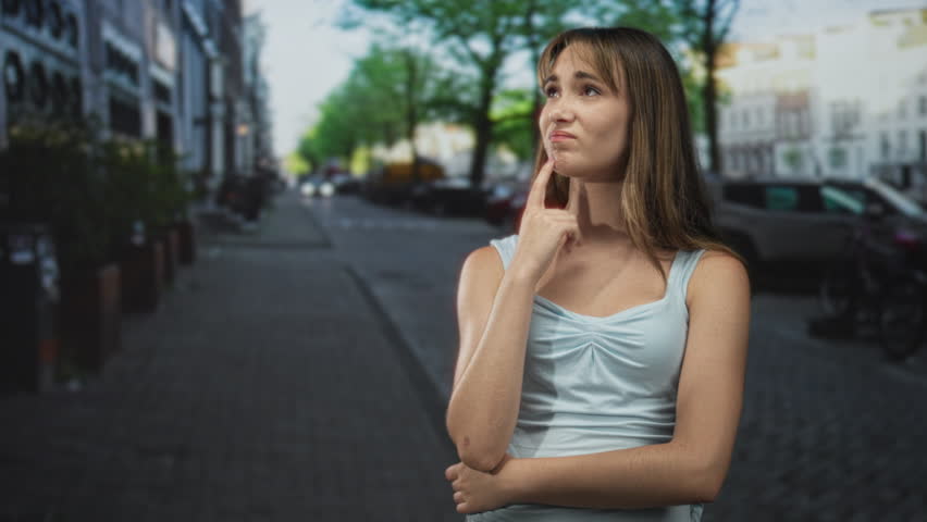 Young woman with finger to chin looking up on a cobblestone city street beside parked cars and trees; contemplation doubt.