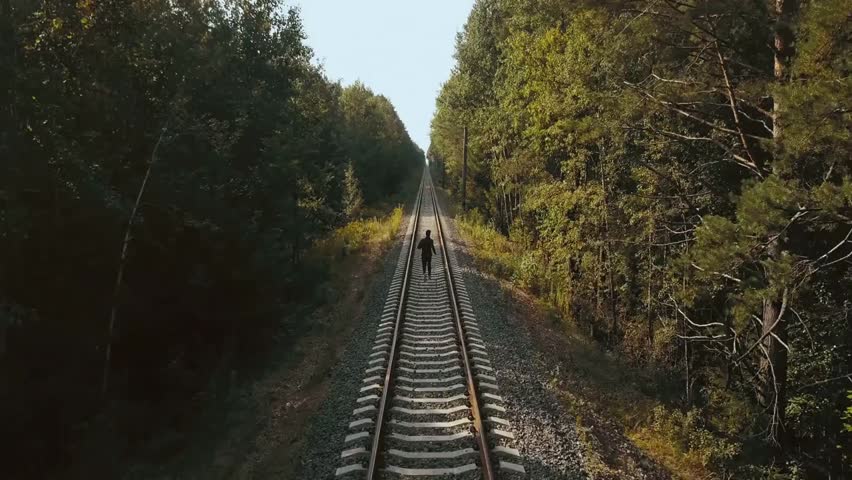 Drone back view of a man running between rail lines through an autumn forest, capturing athletic motion and adventure on the railroad.