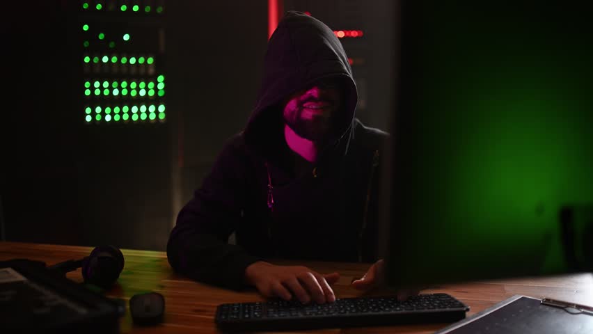 Anonymous hacker in a black hoodie coding in a dark server room. Cybercriminal committing internet crime and data theft with a smile