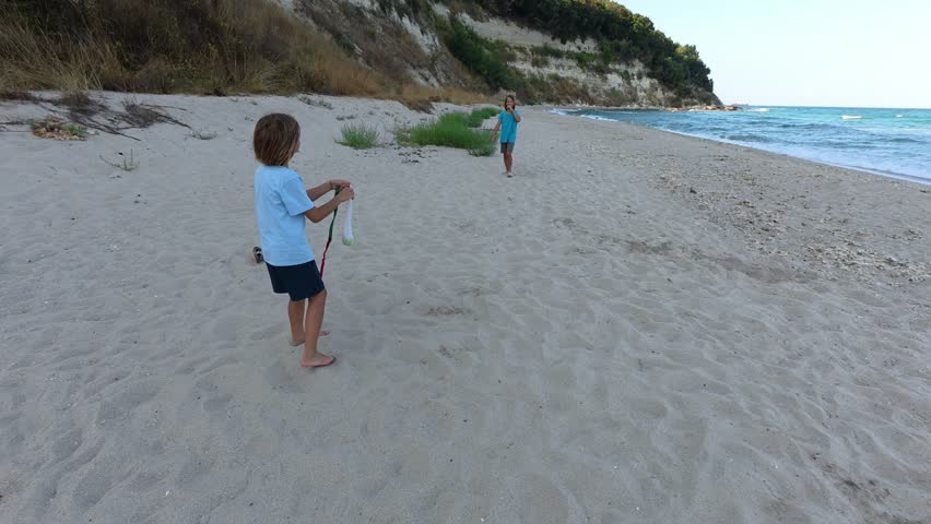 Two children playing catch on sandy beach near sea cliffs. Carefree summer game with flying ball, seaside nature and joyful childhood atmosphere by the ocean