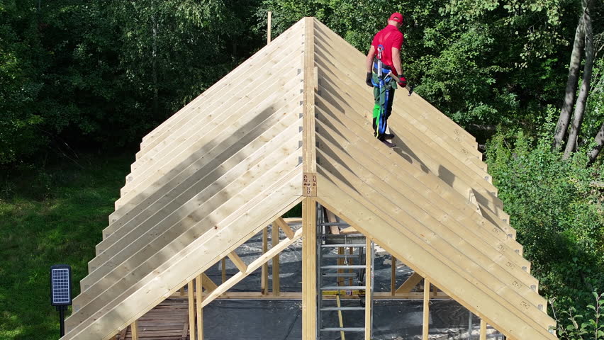 A person works on a wooden roof of a structure in the woods during daylight.