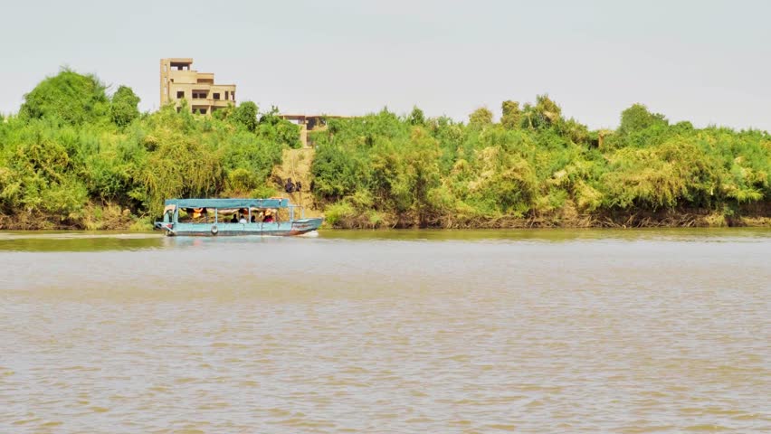 Aerial follow shot of a boat moving along the Nile River near Khartoum, Sudan, capturing calm river flow, surrounding shoreline, and natural African landscape under daylight. The scene reflects transportation, daily life, and iconic river geography. Filmed on 24 December 2025.