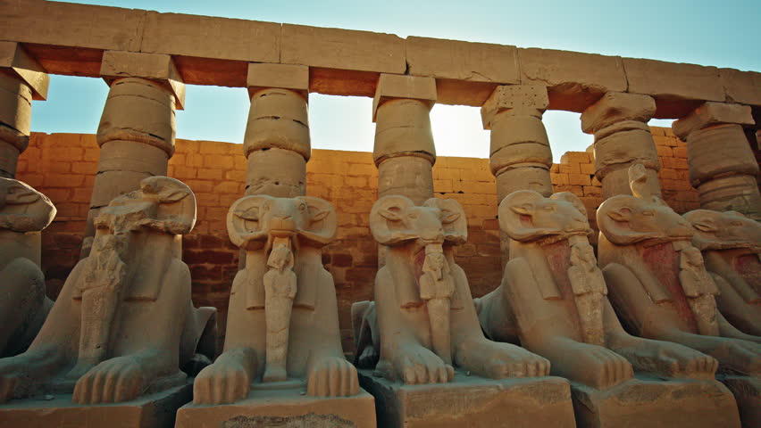 Row of ancient ram headed sphinx statues aligned in front of massive stone columns inside the Karnak Temple, illuminated by warm sunlight, representing sacred architecture and ancient Egyptian
