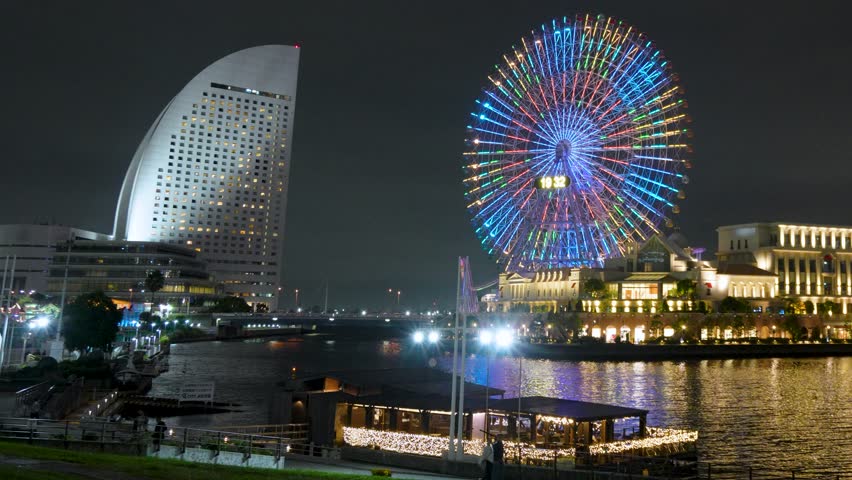 Vibrant autumn night view of Minato Mirai in Yokohama, Japan. Features the brightly lit Cosmo Clock Ferris wheel, modern skyscrapers, calm waterfront reflections, and glowing city lights.