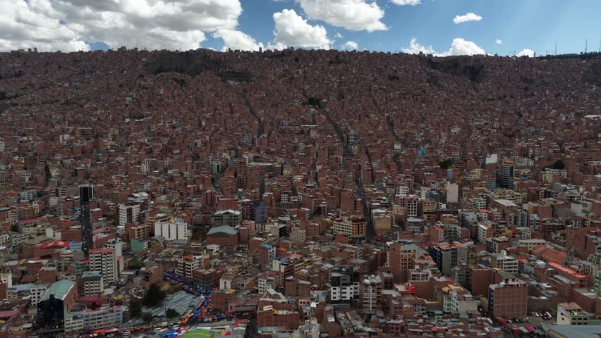 La Paz, Bolivia; March 21, 2025:
This aerial photograph reveals an immense high-altitude settlement extending across steep Andean terrain, forming a striking visual pattern of countless structures built closely together. From this elevated perspective, the viewer can appreciate how homes and mid-rise buildings fill every available space, rising from the lower zones and climbing all the way up the rugged hillsides. The dense composition of reddish brick, concrete, metal rooftops, and narrow lanes creates a dramatic mosaic shaped by decades of expansion in a challenging environment.
The scene highlights how construction has adapted to powerful slopes, producing an intricate network of ascending routes that carve through the thick concentration of homes. The visual effect resembles an enormous tapestry, where lines of streets run vertically toward the mountaintop while the buildings sit tightly packed along the inclines. Soft daylight passing through the clouds illuminates sections of the slope, revealing contrasting tones, shadows, and hints of architectural variation within the overwhelming mass of structures.
The mountains surrounding the settlement act as a natural frame, emphasizing the scale and altitude of the region. Their dark rocky formations and elevated ridges add depth and context, showing how communities have developed in harmony with harsh topography. The image conveys resilience, adaptation, and the complexity of living at significant elevation, where construction challenges are met with creativity and determination.
