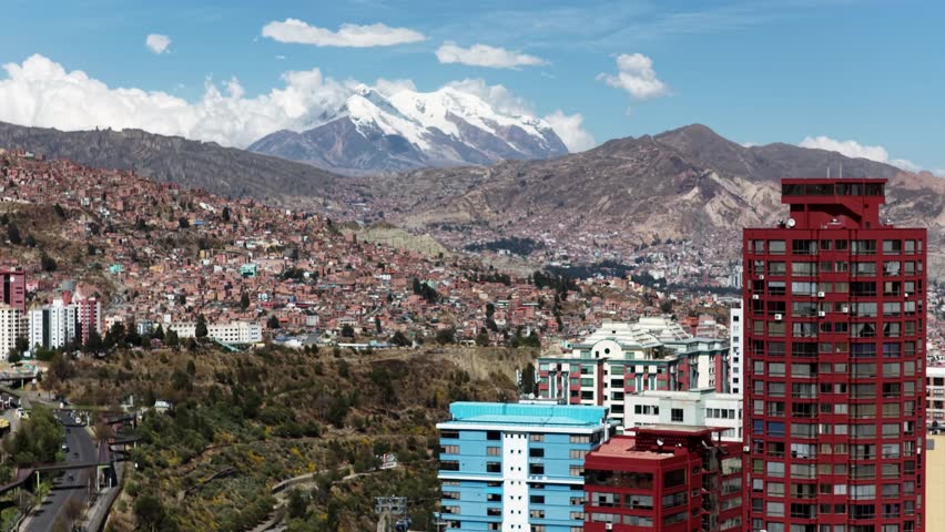 Panoramic urban landscape of La Paz, Bolivia, with Mount Illimani rising majestically in the background. This high-altitude city view captures the dramatic contrast between dense urban development and the rugged Andean mountains. Residential neighborhoods cover the hillsides, while modern buildings and infrastructure fill the valleys, creating a striking visual composition between nature and city life.
Mount Illimani, the iconic snow-capped peak with its three prominent summits, dominates the skyline and serves as a powerful natural symbol of Bolivia. The clear blue sky and bright daylight enhance the textures of the mountains, rocky terrain, and colorful urban architecture. The image reflects the unique geography of La Paz, one of the highest capital cities in the world, where daily life unfolds beneath towering Andean peaks.
This photograph is ideal for travel publications, tourism promotion, geography and culture-related projects, documentaries, editorial use, and educational content. It visually represents themes such as high-altitude living, Andean culture, South American cities, urban expansion, mountain landscapes, and the coexistence of modern architecture with ancient natural formations. Perfect for illustrating destinations in Bolivia, environmental storytelling, and cinematic visual backgrounds.
