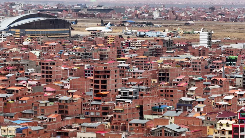 Aerial drone view of El Alto city and El Alto International Airport in Bolivia, showcasing a dense urban landscape at high altitude in the Andes. The clip begins with a wide panoramic view of El Alto’s characteristic red brick buildings, unfinished constructions, and colorful rooftops, revealing the unique architectural style of one of the highest cities in the world. As the drone slowly advances and stabilizes, the scene expands toward the airport area, where commercial airplanes are visible parked on the runway and near hangars, emphasizing the strategic importance of this airport as a major gateway to La Paz and western Bolivia.
The drone maintains a smooth, cinematic movement, creating a sense of scale and depth while highlighting the contrast between the compact urban sprawl and the open airport infrastructure. The surrounding highland terrain and distant neighborhoods reinforce the geographical context of El Alto as a rapidly growing metropolitan area located over 4,000 meters above sea level. This aerial footage captures daily life, transportation, and urban development in a South American city shaped by altitude, migration, and economic activity.
Ideal for documentaries, news reports, travel films, urban studies, aviation content, and socio-economic storytelling, this clip represents modern Bolivia from above. The scene conveys themes of infrastructure, mobility, population density, and city expansion in Latin America, making it suitable for editorial and commercial use related to geography, travel, urbanization, and aviation.