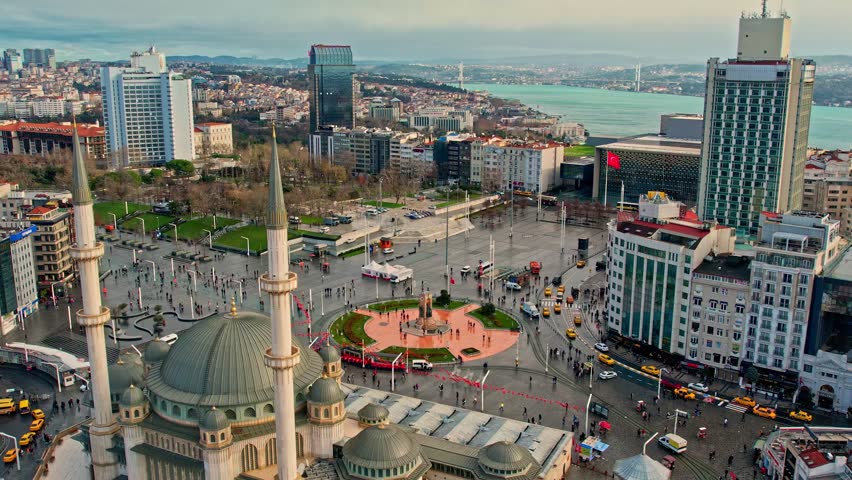 Daytime aerial view of Taksim Square and Taksim Mosque with the Bosphorus Sea in the background. A dynamic Istanbul city center scene combining urban life, coastline, and modern architecture.