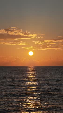 Beautiful orange sun slowly sinking below the horizon over the rippling ocean. Golden sunlight reflects on the tranquil water surface