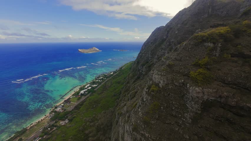 Cinematic aerial drone footage flying along a steep rocky coastal cliff overlooking turquoise ocean waters on Oahu, Hawaii. Scenic tropical coastline with dramatic mountain terrain, clear blue sea, and island shoreline viewed from above. Ideal for travel, nature, adventure, and tourism concepts.