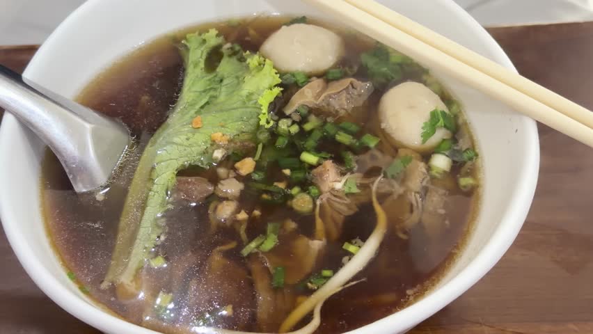 Chili flakes are added to a bowl of Thai pork noodle soup with a spoon