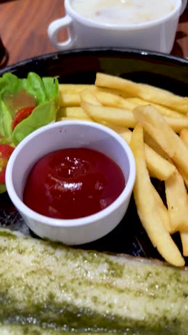 Close-up rotating view of fish steak, french fries, salad, and soup under warm restaurant lighting
