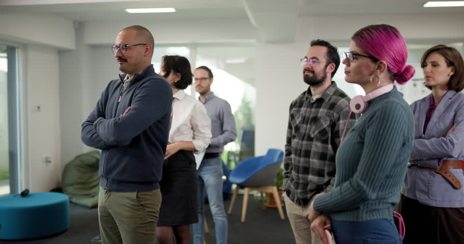 A group of team members stands in an office setting, sharing thoughts and reactions during a meeting. Men and women of different backgrounds interact and exchange ideas.