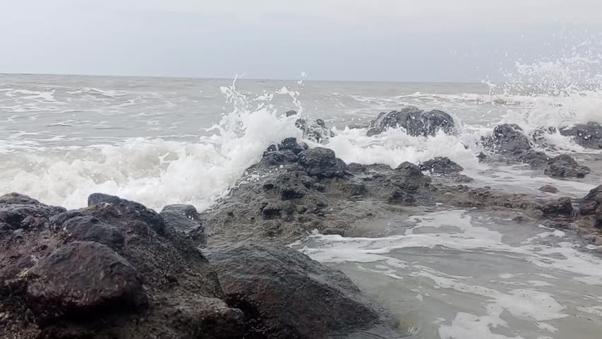 Footage of a natural rocky beach scene with white waves crashing against black coral. A cloudy sky creates a calm, cool, and peaceful seascape